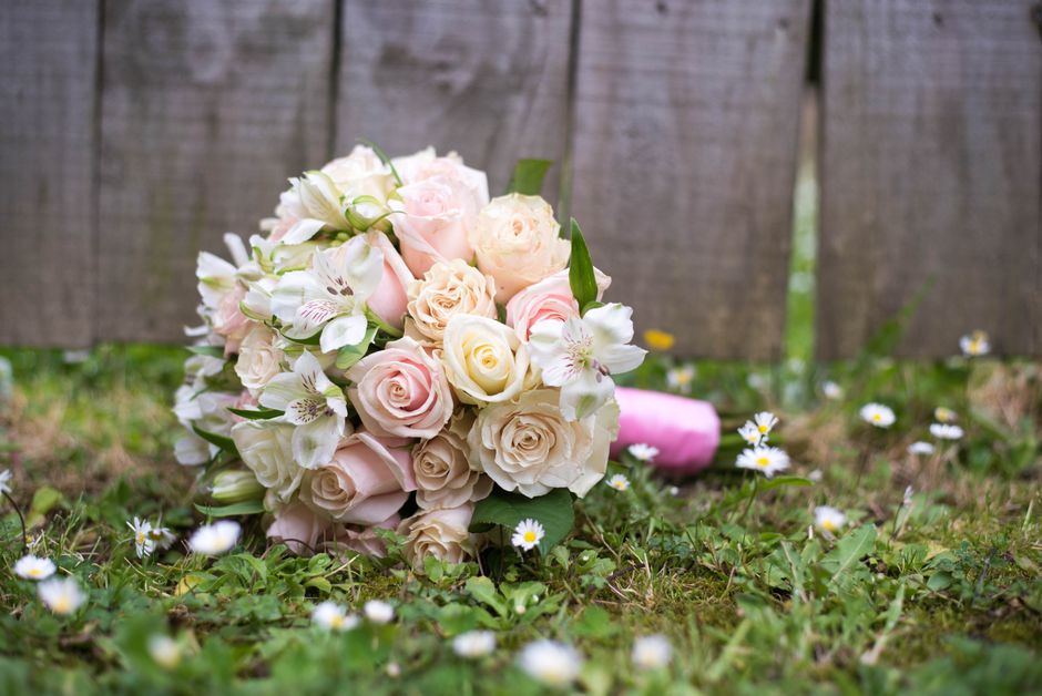 A bouquet of fresh pale pink and cream roses and white lilies with light green foliage, placed on the grass near a weathered wooden fence. The bouquet is positioned at an angle, with some flowers faci