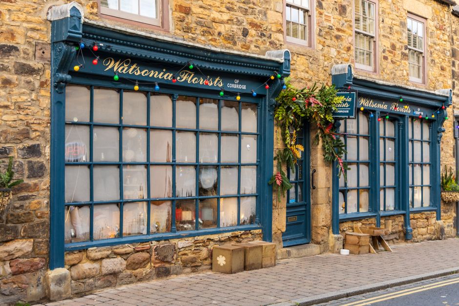 A floral shop storefront with a vintage stone facade and blue wooden framing around large windowpanes covered in frosted glass. The shop, Watsonia Florists of Corbridge, is decorated with colorful str