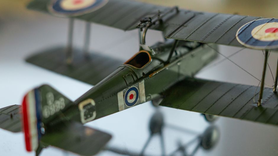 Close-up of a detailed model of a vintage military aircraft with dark green and gray camouflage, featuring roundel insignia and fine panel lines, set in an indoor environment with soft lighting that h