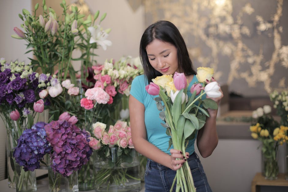 A young woman with shoulder-length dark hair and light skin is holding a bouquet of fresh flowers, including pink tulips, white roses, and yellow freesias, in a flower shop. She is wearing a bright bl